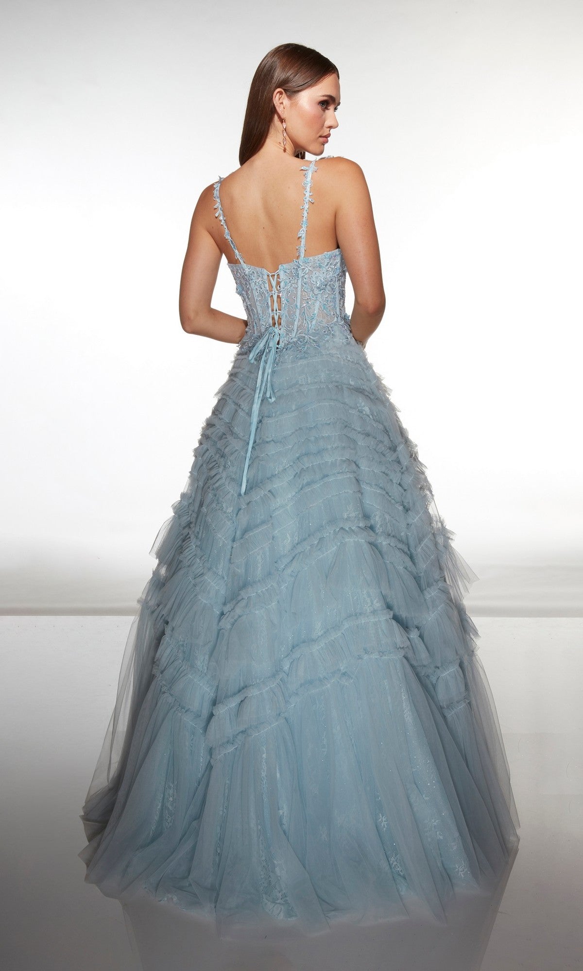 A woman faces away against a plain background wearing the Alyce 62058, a long formal dress in light blue featuring a floor-length ruffled skirt and lace-up back.